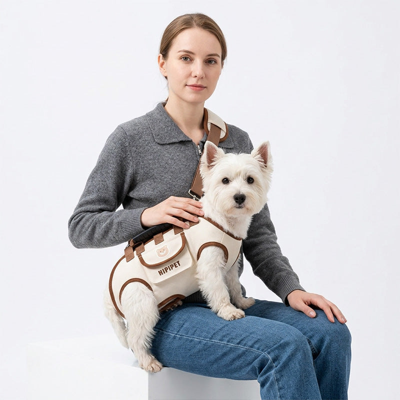 Seated woman wearing a khaki dog carrier harness with a white terrier supported in horizontal carry position.