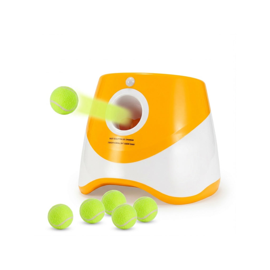 White background studio shot of an orange-and-white automatic dog ball launcher with green tennis balls flying and clustered below.