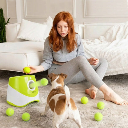 Girl plays with a green automatic dog ball launcher as a small brown-and-white dog chases tennis balls in a bright living room.