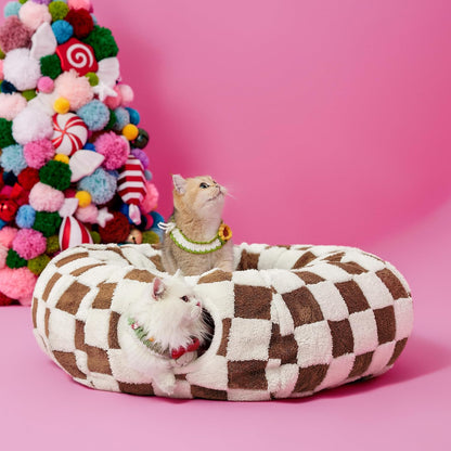 Two cats inside the brown and cream checkered donut cat tunnel bed: one curled in the center cushion, one watching from the tunnel entrance.