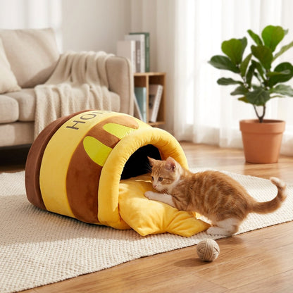 Ginger kitten explores a Honey Pot Cat Cave Bed on a cream rug in a cozy living room with sofa, plant, and wood floor.