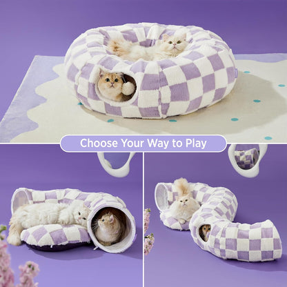Pastel purple checkered cat tunnel bed on a purple studio backdrop with two cats — one on top, one peeking out the tunnel.